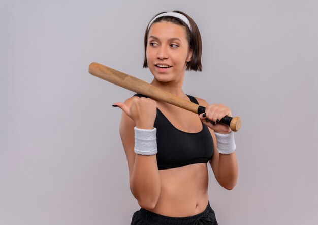 Young fitness woman in sportswear holding baseball bat looking aside with smile on face pointing back with thumb standing over white wall