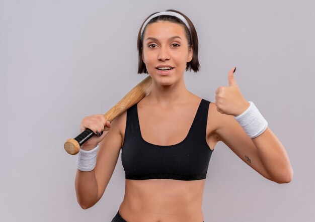 Молодая фитнес-женщина в спортивной одежде, держащая бейсбольную биту, уверенно улыбаясь, показывает палец вверх, стоя над белой стеной