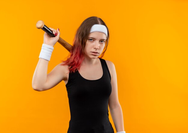 オレンジ色の壁の上に立っている深刻な顔で野球のバットを保持しているスポーツウェアの若いフィットネス女性