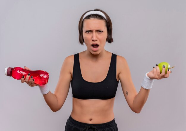 Young fitness woman in sportswear holding bottle of water and green apple shouting with angry face standing over white wall