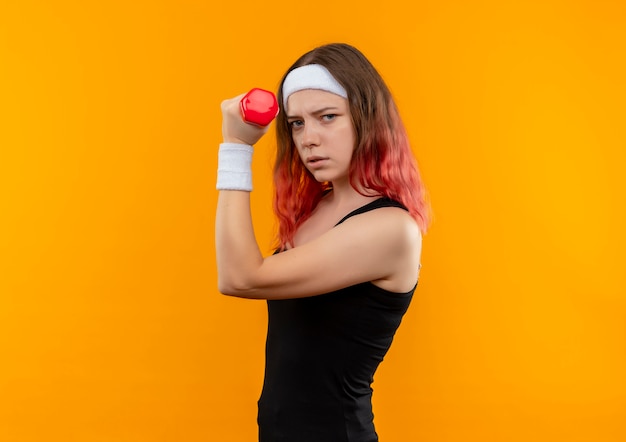 オレンジ色の壁の上に立っている深刻な顔でパワーエクササイズを行うダンベルを保持しているスポーツウェアの若いフィットネス女性