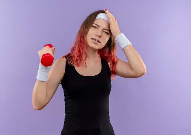 Young fitness woman in sportswear holding dumbbell looking tired standing over purple wall