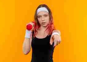 Free photo young fitness woman in sportswear holding dumbbell pointing with index finger to camera with saerious face standing over orange wall