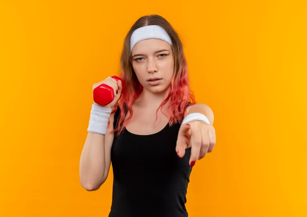 Young fitness woman in sportswear holding dumbbell pointing with index finger to camera with saerious face standing over orange wall