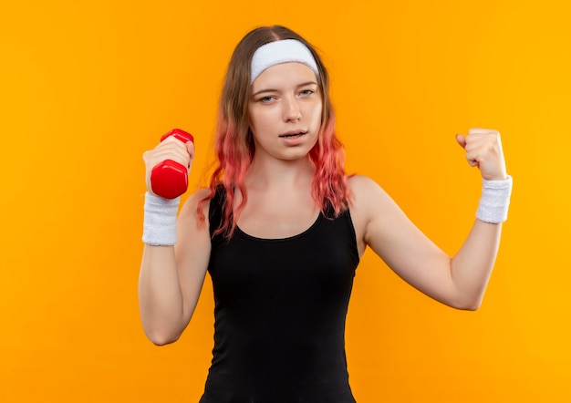 オレンジ色の壁の上に立っている勝者のように拳を上げるダンベルを保持しているスポーツウェアの若いフィットネス女性