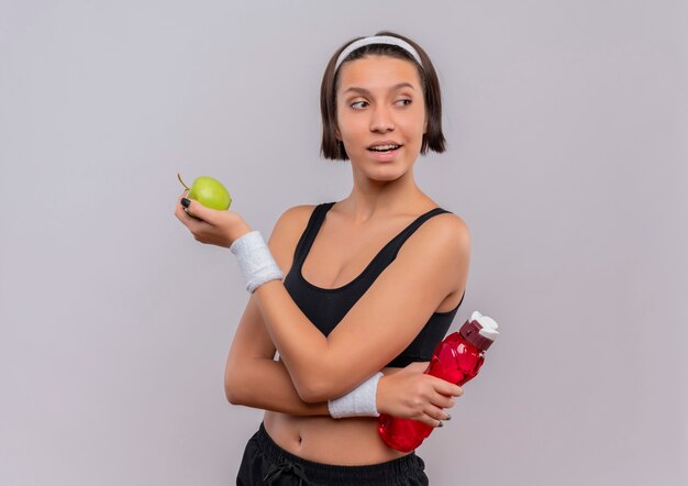 青リンゴと水のボトルを保持しているスポーツウェアの若いフィットネス女性は、白い壁の上に立っている自信を持って笑顔で脇を見て
