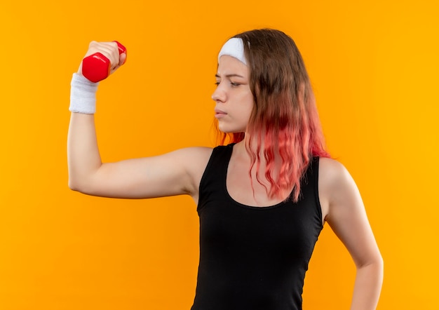Young fitness woman in sportswear holding holding dumbbell doing exercises with serious face standing over orange wall
