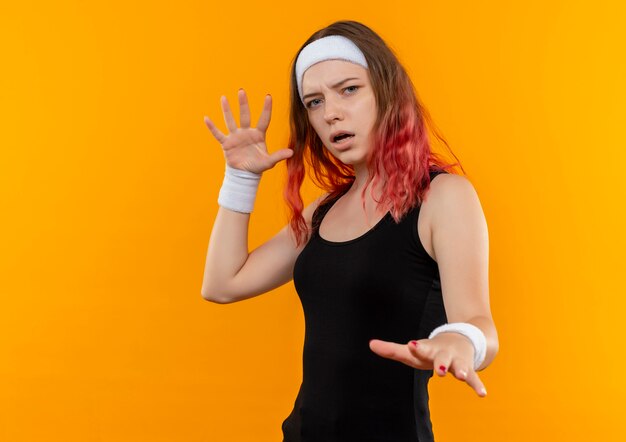 オレンジ色の壁の上に立っている恐怖の表情で防衛ジェスチャーを作る手のひらを保持しているスポーツウェアの若いフィットネス女性