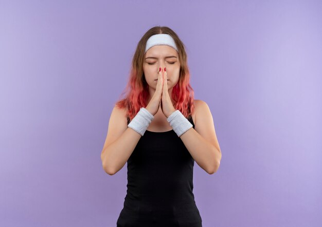 Young fitness woman in sportswear holding palms together like praying with closed eyes with hope expression standing over purple wall