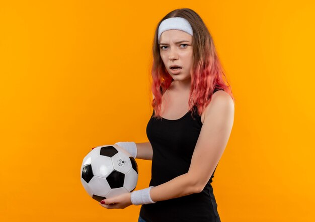 オレンジ色の壁の上に立って不機嫌と混乱してサッカーボールを保持しているスポーツウェアの若いフィットネス女性
