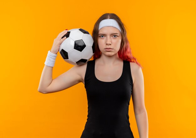 Молодая женщина фитнеса в спортивной одежде держит футбольный мяч в руке с серьезным лицом, стоящим над оранжевой стеной