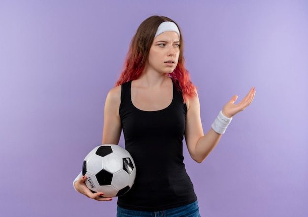 Молодая фитнес-женщина в спортивной одежде держит футбольный мяч в замешательстве, стоя над фиолетовой стеной