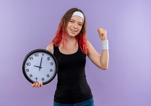 Young fitness woman in sportswear holding wall clock clenching fist happy and exited standing over purple wall