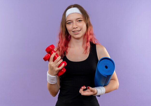 Young fitness woman in sportswear holding yoga mat and dumbbells smiling with happy face standing over purple wall