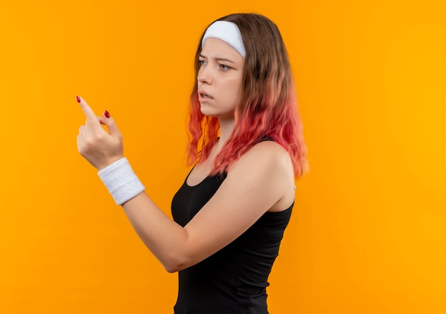 Молодая фитнес-женщина в спортивной одежде смотрит в сторону, указывая пальцем с растерянным выражением лица, стоя над оранжевой стеной