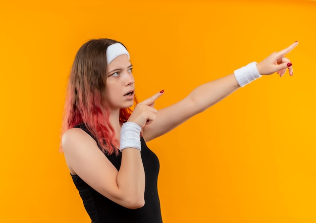 Young fitness woman in sportswear looking aside surprised pointing with fingers of both hands to something standing over orange wall