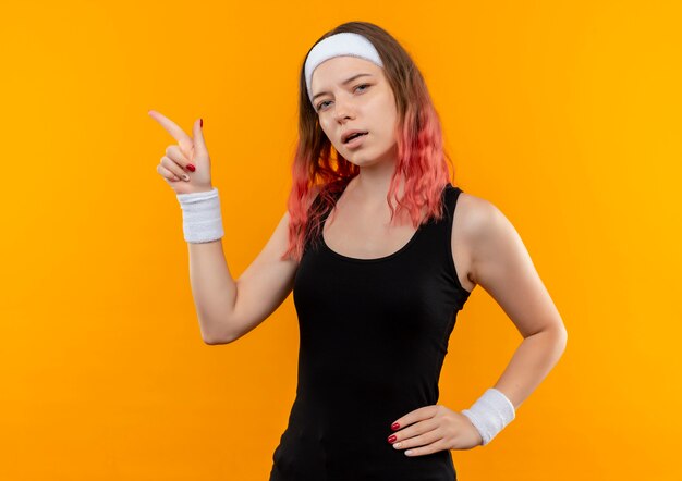 Молодая фитнес-женщина в спортивной одежде выглядит уверенно, указывая пальцем в сторону, стоящую над оранжевой стеной