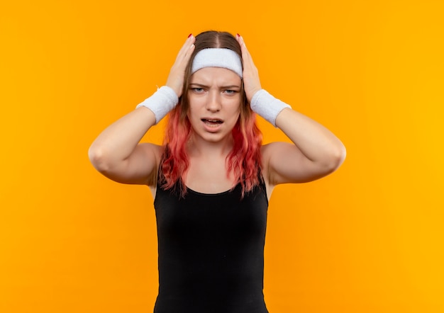 Young fitness woman in sportswear looking confused and very disappointed touching her head standing over orange wall