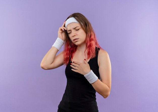 Young fitness woman in sportswear looking unwell and touching head having headache standing over purple wall