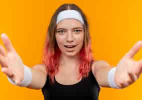 Free photo young fitness woman in sportswear making welcoming gesture with hands standing over orange wall