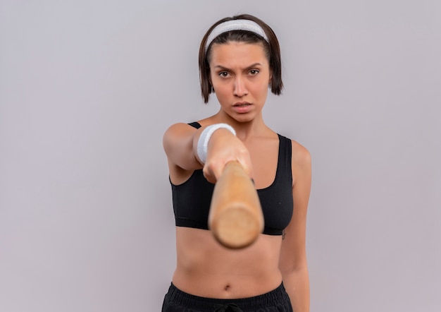Young fitness woman in sportswear pointing with baseball bat to camera with serious face standing over white wall