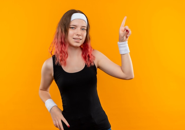 Young fitness woman in sportswear pointing with finger to the side standing over orange wall