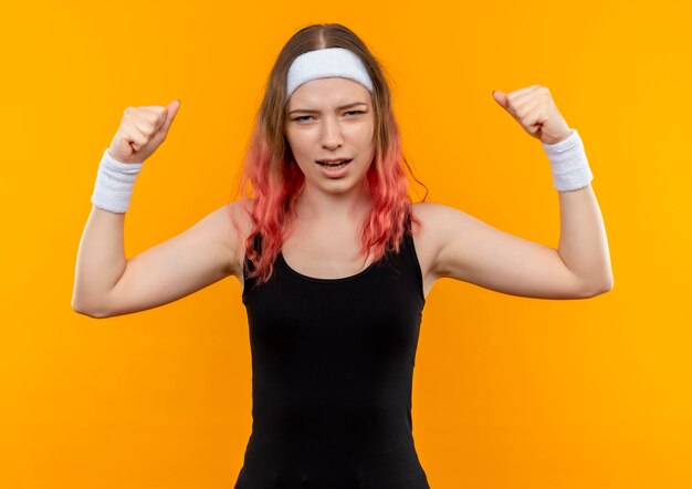 オレンジ色の壁の上に立っているイライラした表情で拳を上げるスポーツウェアの若いフィットネス女性