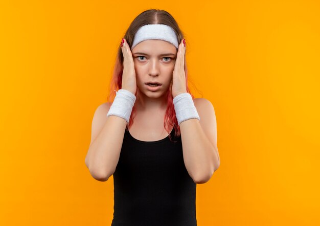 Young fitness woman in sportswear shocked touching her face with arms standing over orange wall