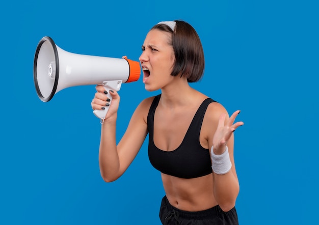 Young fitness woman in sportswear shouting to megaphone with aggressive expression standing over blue wall