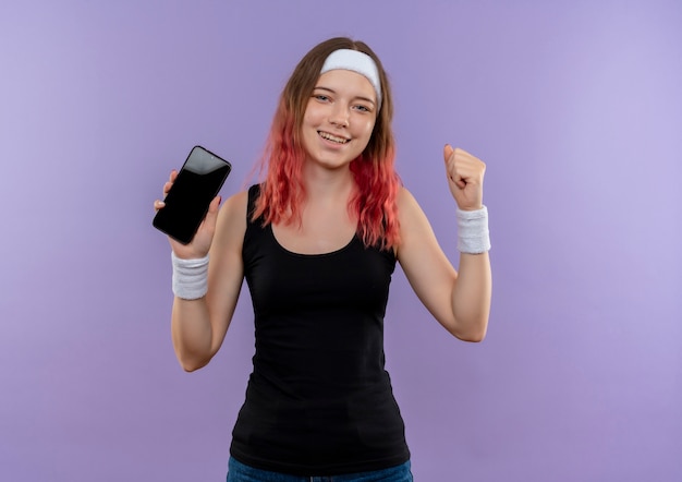 Молодая фитнес-женщина в спортивной одежде показывает смартфон, сжимающий кулак, весело улыбаясь и выходит, стоя над фиолетовой стеной