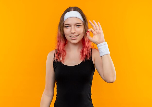 운동복에 젊은 피트 니스 여자 오렌지 벽 위에 서 확인 기호를 유쾌하게 보여주는 미소