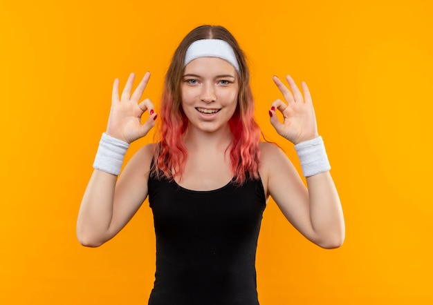 Молодая фитнес-женщина в спортивной одежде, весело улыбаясь, показывая хорошие знаки, обеими руками стоя над оранжевой стеной