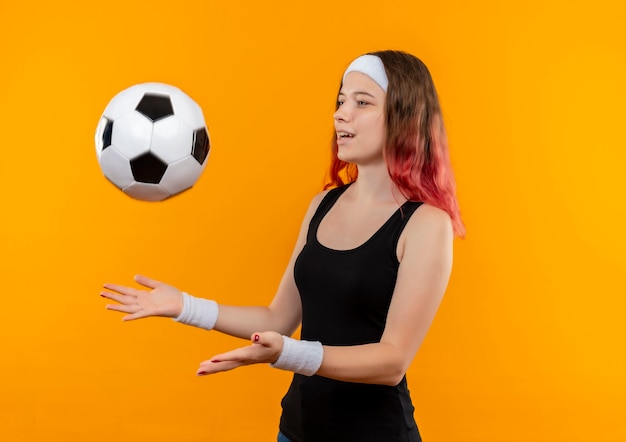 Молодая фитнес-женщина в спортивной одежде, бросая футбольный мяч, счастливая и позитивная, стоит над оранжевой стеной