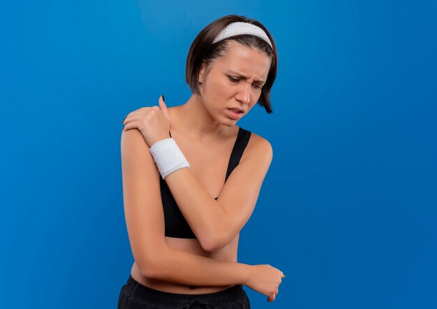 Young fitness woman in sportswear touching her shoulder looking unwell feeling pain standing over blue wall