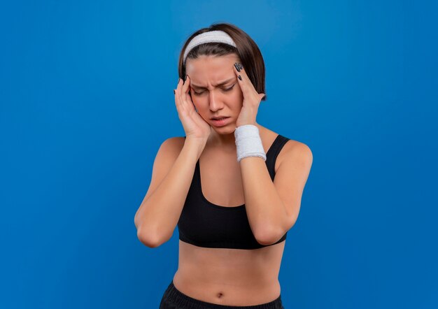 Young fitness woman in sportswear touching her temples having headache standing over blue wall