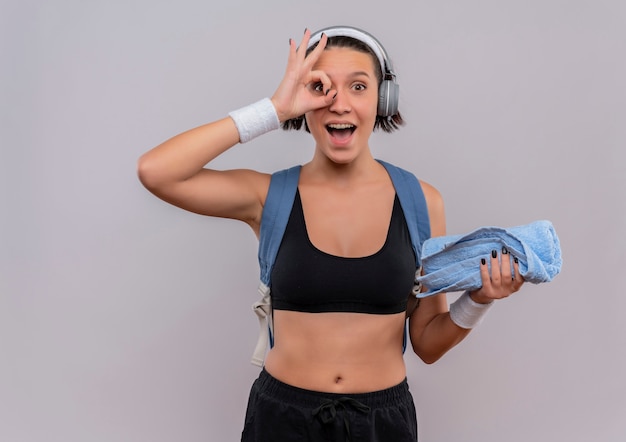 Young fitness woman in sportswear with backpack and headphones on head holding towel smiling cheerfully