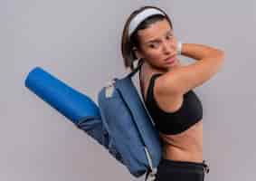 Free photo young fitness woman in sportswear with backpack and yoga mat looking aside displeased and confused standing over white wall