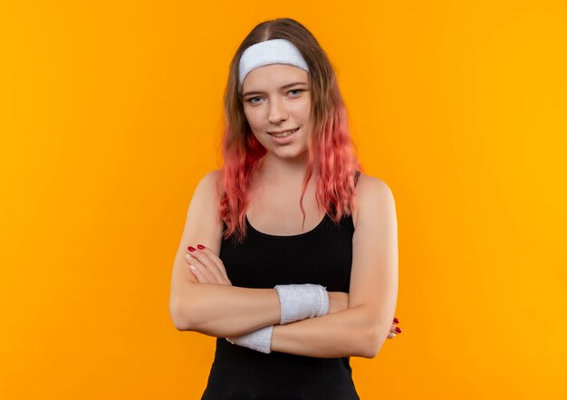 Молодая фитнес-женщина в спортивной одежде со скрещенными руками на груди с уверенной улыбкой на лице, стоящей над оранжевой стеной