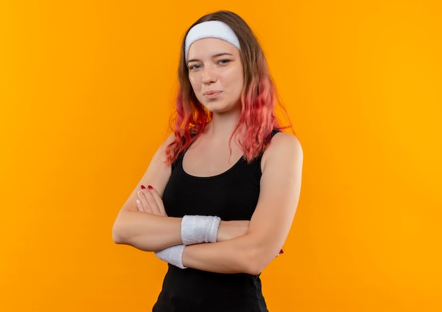 Молодая фитнес-женщина в спортивной одежде со скрещенными руками с уверенной улыбкой на лице, стоящей над оранжевой стеной