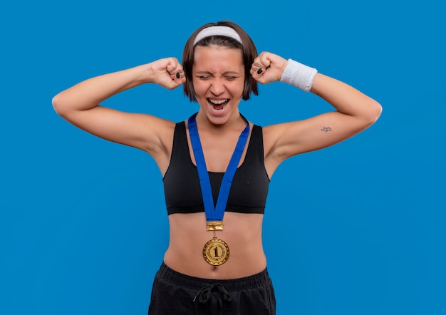 Free photo young fitness woman in sportswear with gold medal around her neck clenching fists rejoicing her success standing over blue wall