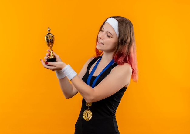 Young fitness woman in sportswear with gold medal around her neck holding trophy looking at it smiling cheerfully standing over orange wall