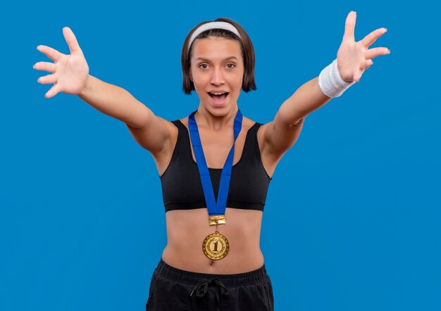 Foto gratuita giovane donna fitness in abbigliamento sportivo con medaglia d'oro al collo che fa gesto di benvenuto ampia apertura mani in piedi sopra la parete blu