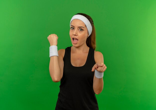 Foto gratuita giovane donna fitness in abiti sportivi con fascia pugno di serraggio puntato con il dito alla telecamera guardando sorpreso in piedi sopra la parete verde