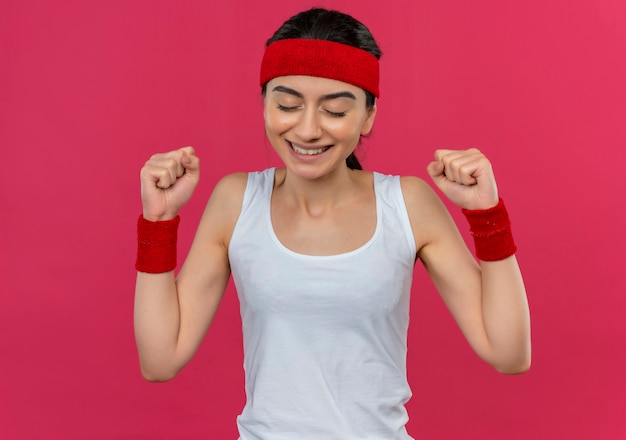 Young fitness woman in sportswear with headband clenching fists happy and excited rejoicing her success standing over pink wall