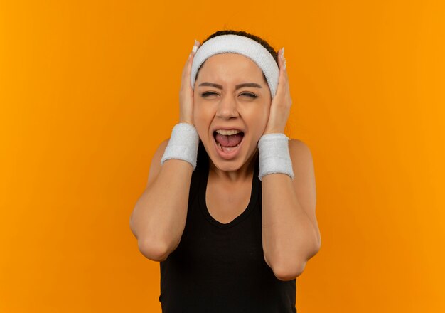 Молодая фитнес-женщина в спортивной одежде с головной повязкой, закрывающей уши руками, кричит с раздраженным выражением лица, стоя над оранжевой стеной