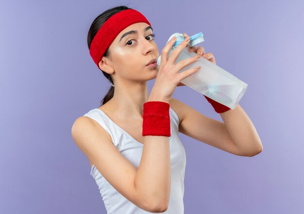 Young fitness woman in sportswear with headband drinking water looking tired standing over purple wall