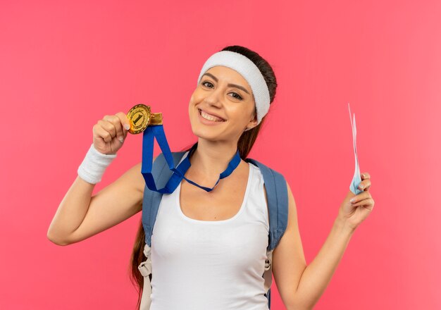 Молодая фитнес-женщина в спортивной одежде с повязкой на голову и золотой медалью на шее с рюкзаком, держащим авиабилеты, весело улыбаясь, стоя над розовой стеной