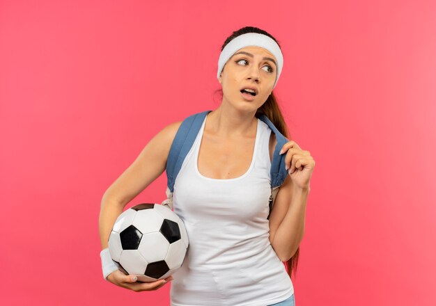 Young fitness woman in sportswear with headband and gold medal around her neck with backpack holding soccer ball looking aside puzzled standing over pink wall