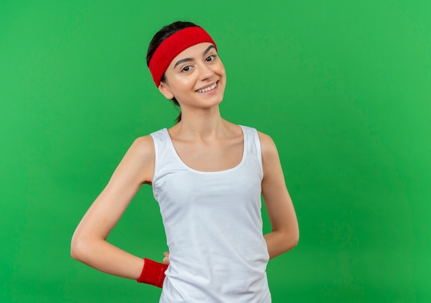 Young fitness woman in sportswear with headband happy and positive smiling cheerfully standing over green wall