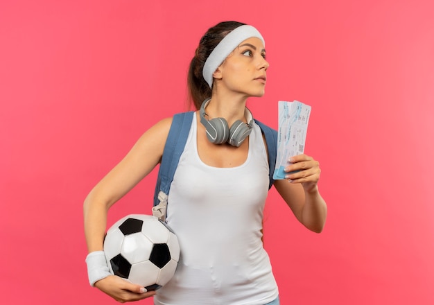 Foto gratuita giovane donna di forma fisica in abbigliamento sportivo con la fascia e le cuffie intorno al suo collo che tiene i biglietti aerei e il pallone da calcio che osserva da parte con la faccia seria che sta sopra il muro rosa